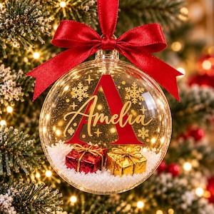 May include: A clear spherical Christmas ornament featuring a large red letter A and the name Amelia in gold script. Inside the ornament are artificial snow, a red gift box, and a gold gift box. A red satin ribbon is tied at the top.