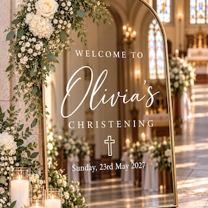 May include: An arched gold-framed mirror sign with white text reading WELCOME TO Olivia's CHRISTENING, a cross icon, and Sunday, 23rd May 2027. The mirror is decorated with white roses, small white flowers, and green eucalyptus leaves.