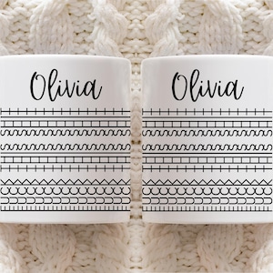 Può includere: Due tazze in ceramica bianca con il nome "Olivia" in corsivo nero. Ogni tazza presenta motivi geometrici neri. Le tazze sono esposte su una superficie bianca a maglia.