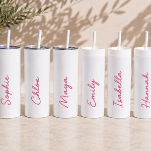 Op de afbeelding: Zes witte cilindrische bekers met deksels en rietjes, elk voorzien van een unieke naam in een roze scriptlettertype. De namen op de bekers zijn Sophie, Chloe, Maya, Emily, Isabella en Hannah. De bekers staan in een rij opgesteld.