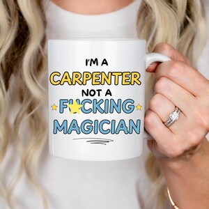 May include: White ceramic mug with the text "I'm a Carpenter not a *F*ucking Magician" in yellow, blue, and black lettering with two yellow stars.