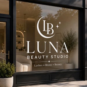 May include: A white vinyl decal applied to a glass storefront window featuring the text LB LUNA BEAUTY STUDIO with a crescent moon and star icons. Below the main text, the words Lashes, Brows, and Beauty are separated by small dots.
