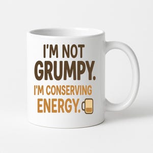 Op de afbeelding: Witte keramische koffiemok met de tekst "I'M NOT GRUMPY. I'M CONSERVING ENERGY." in bruine en beige letters. Naast het woord "ENERGY" staat een klein koffiekopje-pictogram. De mok heeft een gebogen handvat.