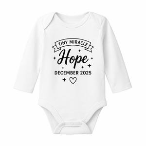 May include: White baby onesie with long sleeves. The onesie features the text "TINY MIRACLE Hope DECEMBER 2025" in black script, with star and heart accents. A sweet and sentimental design for a baby.