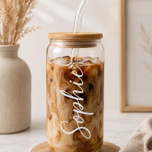 May include: A clear glass tumbler with iced coffee and a glass straw. The tumbler has a bamboo lid and the name "Sophie" in white script. The drink sits on a wooden coaster. A vase and framed art are in the background.