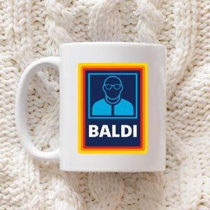 Puede incluir: Taza de cerámica blanca con un gráfico azul y blanco de una persona con gafas dentro de un cuadrado con bordes rojos, amarillos y azules. La palabra "BALDI" está impresa en blanco debajo. La taza tiene un asa blanca.