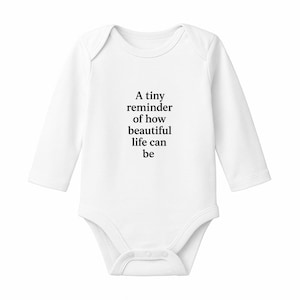 May include: White long-sleeve baby bodysuit with the text "A tiny reminder of how beautiful life can be" in black. The bodysuit has a round neckline and snap closures at the bottom.