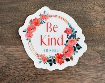 May include: A white sticker with a floral wreath design. The wreath is composed of red and pink flowers with green leaves. The text "Be Kind Of A Bitch" is in the center, with "Be" in white, "Kind" in red, and "Of A Bitch" in black.