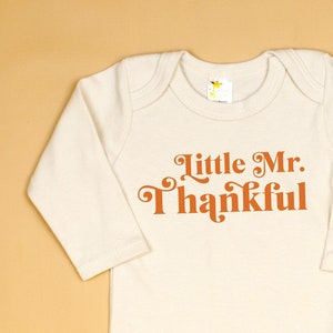 Peut inclure: Un body bébé à manches longues de couleur crème avec le texte "Little Mr. Thankful" en orange.