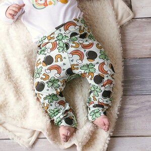 May include: A baby wearing a white onesie with the text "My 1st Patrick's Day!" and green and white patterned pants with shamrocks, rainbows, and gold pots of gold. The baby is lying on a white fluffy blanket.