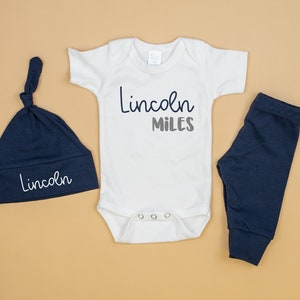 May include: A white baby onesie with the text "Lincoln Miles" in gray and white lettering. A navy blue hat with the text "Lincoln" in white lettering. A pair of navy blue baby leggings.
