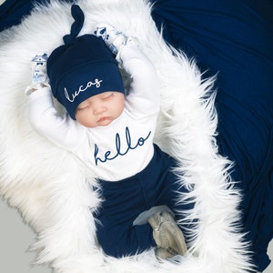 May include: A newborn baby wearing a navy blue and white outfit with the word "hello" on the front. The baby is wearing a navy blue hat with the name "Lucas" embroidered on it. The baby is lying on a white fluffy blanket.