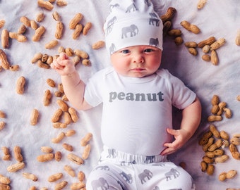 Könnte beinhalten: Ein Baby trägt einen weißen Strampler mit dem Wort "Peanut" darauf. Das Baby trägt eine graue und weiße Mütze mit Elefanten darauf. Das Baby liegt auf einer weißen Decke, umgeben von Erdnüssen.