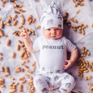 May include: A baby wearing a white onesie with the word "peanut" printed on it. The baby is wearing a gray and white hat with elephants printed on it. The baby is lying on a white blanket surrounded by peanuts.