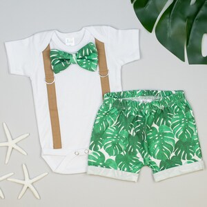 May include: A white bodysuit with brown suspenders and a green bow tie with a tropical leaf pattern. The outfit is paired with green shorts featuring a tropical leaf print.