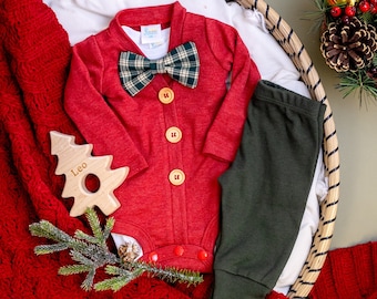 Puede incluir: Un mono rojo para bebé con una pajarita a cuadros, pantalones verdes, un juguete de árbol de Navidad de madera y una manta de punto roja. El mono tiene botones en la parte delantera y el juguete tiene el nombre "Leo" grabado.