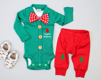 Puede incluir: Un cárdigan verde con una pajarita roja y blanca, un pantalón rojo con árboles verdes y un par de zapatos blancos con árboles rojos y verdes. El cárdigan tiene el texto "my first Christmas" bordado.