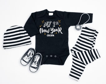 May include: Black and white striped baby outfit with a black long-sleeved onesie that says "My 1st New Year Colton" in gold lettering. The outfit also includes a black and white striped hat and pants.