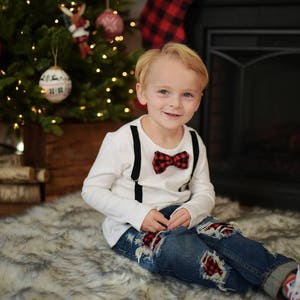 May include: A young boy wearing a white long-sleeved shirt with black suspenders and a red and black bow tie. He is wearing blue jeans with red and black patches and red and white sneakers. He is sitting on a white furry rug in front of a fireplace.