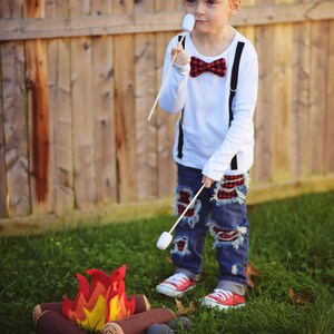 May include: A young boy wearing a white shirt with a red and black bow tie and blue jeans with red plaid patches is roasting a marshmallow over a felt campfire. The boy is holding a wooden stick with a marshmallow on the end. The campfire is made of felt logs and has a felt flame.