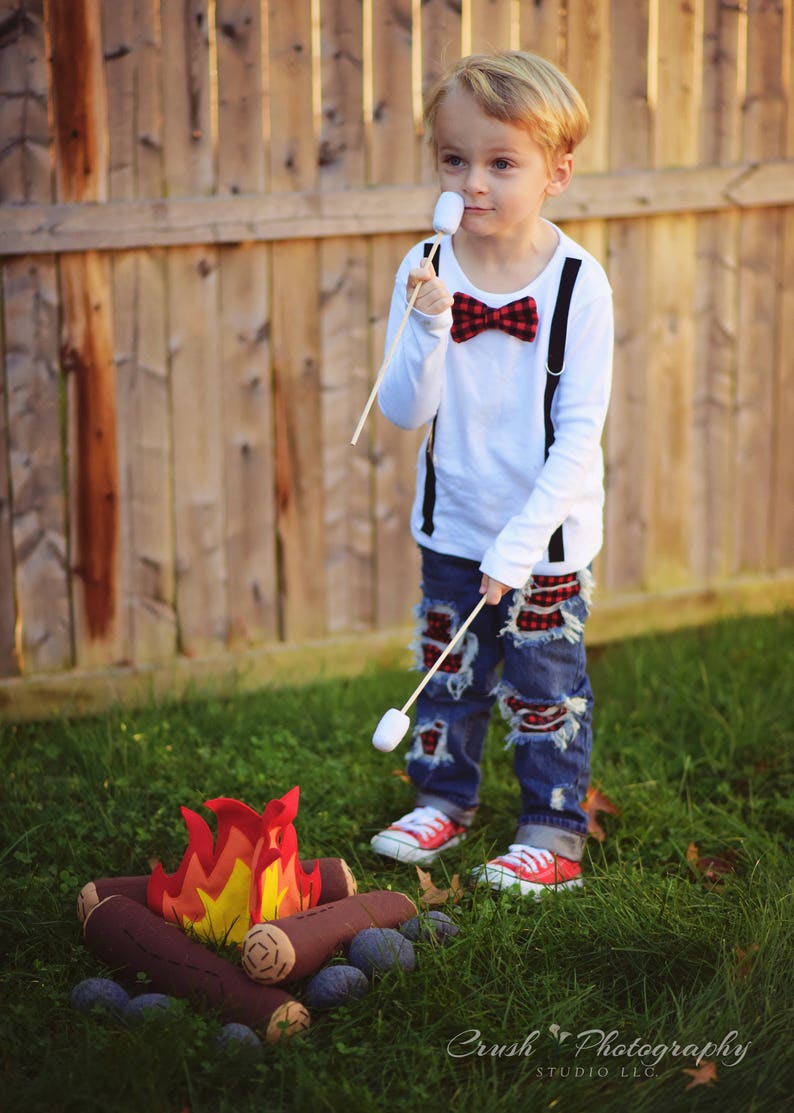 May include: A young boy wearing a white shirt with a red and black bow tie and blue jeans with red plaid patches is roasting a marshmallow over a felt campfire. The boy is holding a wooden stick with a marshmallow on the end. The campfire is made of felt logs and has a felt flame.