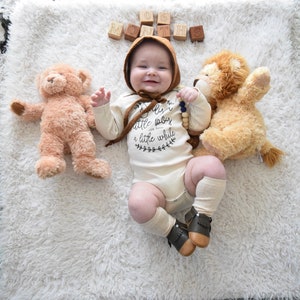 Pode incluir: Um beb&ecirc; vestindo um macac&atilde;o branco com o texto "Eu sou apenas um beb&ecirc; pequeno por um tempo" e um chap&eacute;u marrom est&aacute; deitado em um tapete branco e felpudo com dois animais de pel&uacute;cia. O beb&ecirc; est&aacute; sorrindo e segurando um colar de contas de madeira. H&aacute; blocos de madeira que soletram "SEIS MESES" no tapete.