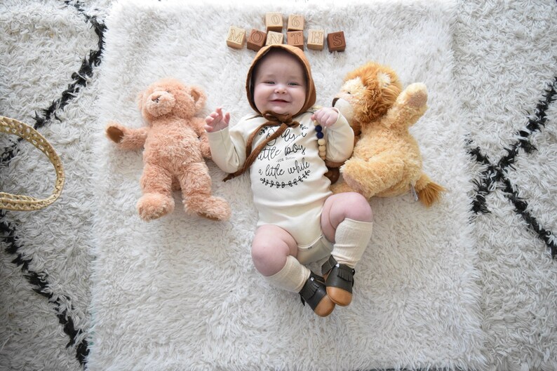 Pode incluir: Um beb&ecirc; vestindo um macac&atilde;o branco com o texto "Eu sou apenas um beb&ecirc; pequeno por um tempo" e um chap&eacute;u marrom est&aacute; deitado em um tapete branco e felpudo com dois animais de pel&uacute;cia. O beb&ecirc; est&aacute; sorrindo e segurando um colar de contas de madeira. H&aacute; blocos de madeira que soletram "SEIS MESES" no tapete.