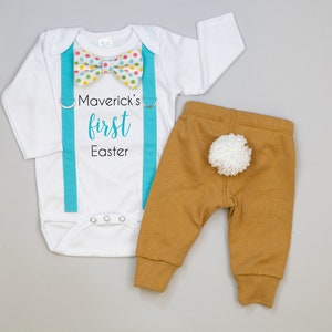Puede incluir: Mono blanco de manga larga con tirantes turquesa y una pajarita de lunares multicolores. El mono tiene el texto "Maverick's first Easter" impreso. Pantal&oacute;n marr&oacute;n con una cola de pomp&oacute;n blanco.