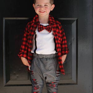May include: A young boy wearing a red and black plaid shirt, a white undershirt, a red bow tie, gray ripped jeans, and black suspenders.