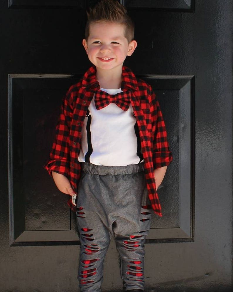 May include: A young boy wearing a red and black plaid shirt, a white undershirt, a red bow tie, gray ripped jeans, and black suspenders.