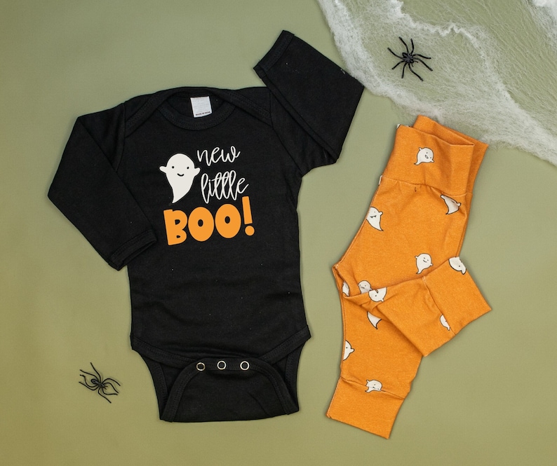Pu&ograve; includere: Body nero a maniche lunghe con il testo "new little BOO!" e una grafica di un fantasma. Pantaloni arancioni con un motivo a fantasma.