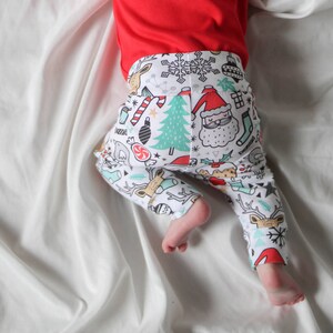 May include: A baby wearing white and red Christmas-themed leggings with a variety of holiday-themed illustrations, including reindeer, candy canes, snowflakes, and Santa Claus. The leggings are on a white background.