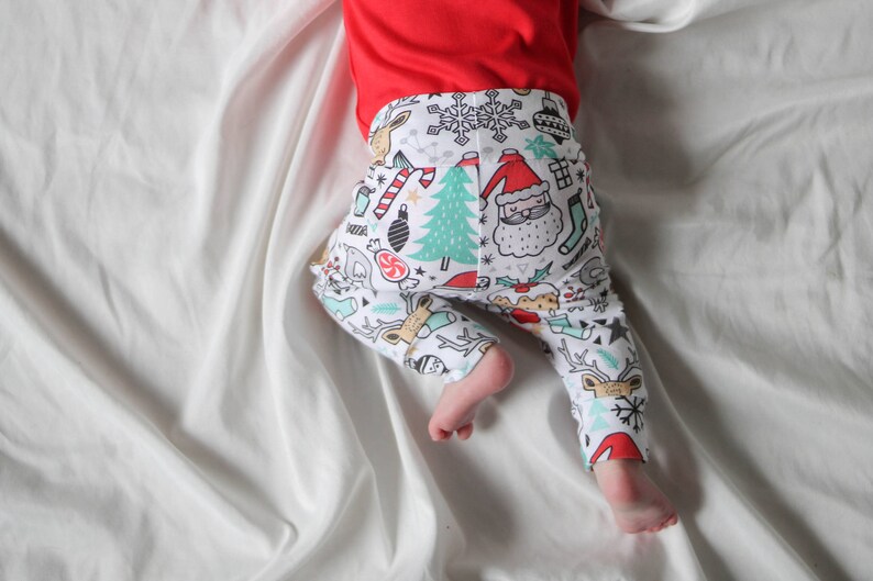 May include: A baby wearing white and red Christmas-themed leggings with a variety of holiday-themed illustrations, including reindeer, candy canes, snowflakes, and Santa Claus. The leggings are on a white background.
