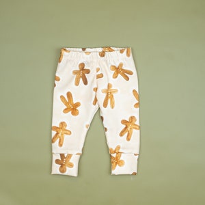 Puede incluir: Leggings de beb&eacute; blancos con estampado de hombre de jengibre. Los leggings tienen un estampado de hombre de jengibre marr&oacute;n sobre un fondo blanco.