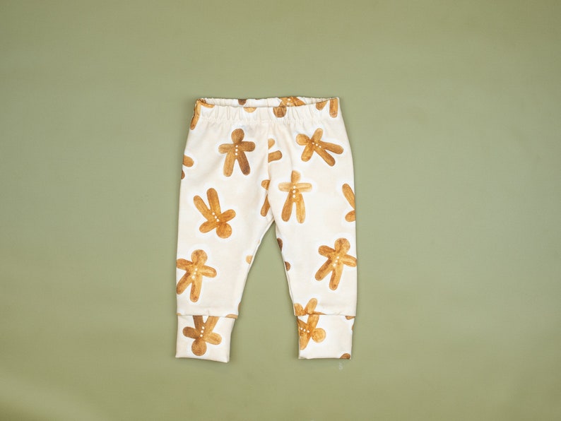 Puede incluir: Leggings de beb&eacute; blancos con estampado de hombre de jengibre. Los leggings tienen un estampado de hombre de jengibre marr&oacute;n sobre un fondo blanco.