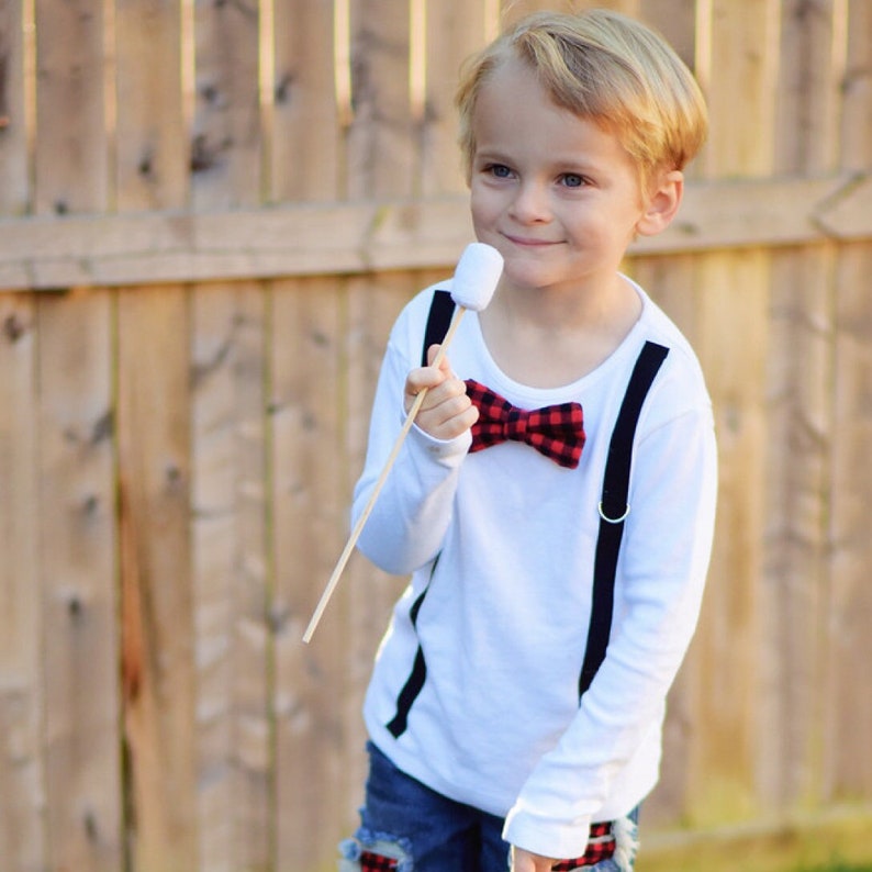 May include: A young boy wearing a white long-sleeved shirt with black suspenders and a red and black bow tie holds a stick with a marshmallow on it.