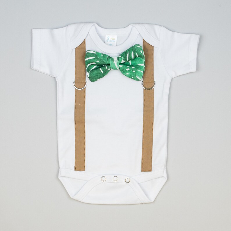 May include: A white baby onesie with brown suspenders and a green bow tie with a tropical leaf pattern.