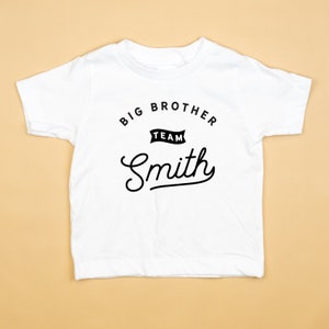 Puede incluir: Camiseta blanca con texto negro que dice "Big Brother Team Smith" en una fuente de estilo vintage.
