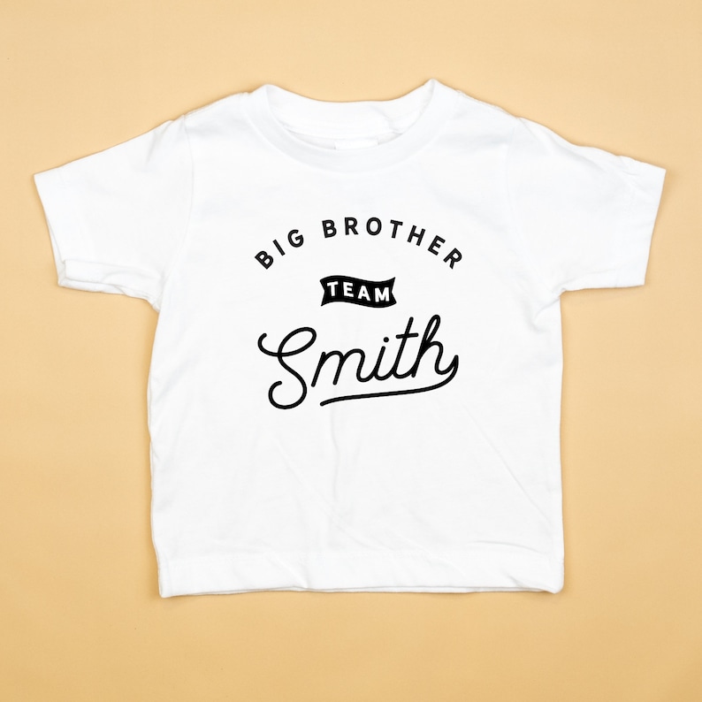 Puede incluir: Camiseta blanca con texto negro que dice "Big Brother Team Smith" en una fuente de estilo vintage.