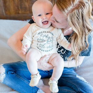 Pode incluir: Uma mulher est&aacute; segurando um beb&ecirc; que est&aacute; vestindo um macac&atilde;o branco com o texto "Only a little boy for such a little while". O beb&ecirc; est&aacute; usando sapatos cor de creme. A mulher est&aacute; usando jeans azul e uma camisa cinza.