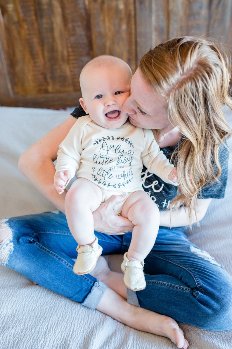 Pode incluir: Uma mulher est&aacute; segurando um beb&ecirc; que est&aacute; vestindo um macac&atilde;o branco com o texto "Only a little boy for such a little while". O beb&ecirc; est&aacute; usando sapatos cor de creme. A mulher est&aacute; usando jeans azul e uma camisa cinza.