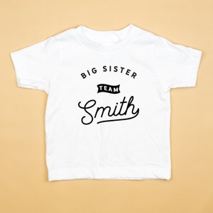 Puede incluir: Camiseta blanca con texto negro que dice "Big Sister Team Smith" en una fuente de escritura.