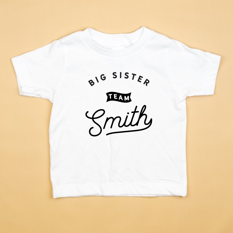 Puede incluir: Camiseta blanca con texto negro que dice "Big Sister Team Smith" en una fuente de escritura.
