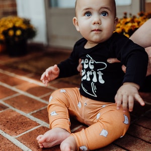 Pu&ograve; includere: Un bambino che indossa una maglietta nera a maniche lunghe con il testo "Free Spirit" e pantaloni arancioni con un motivo a fantasmi. Il bambino &egrave; seduto su un patio in mattoni.