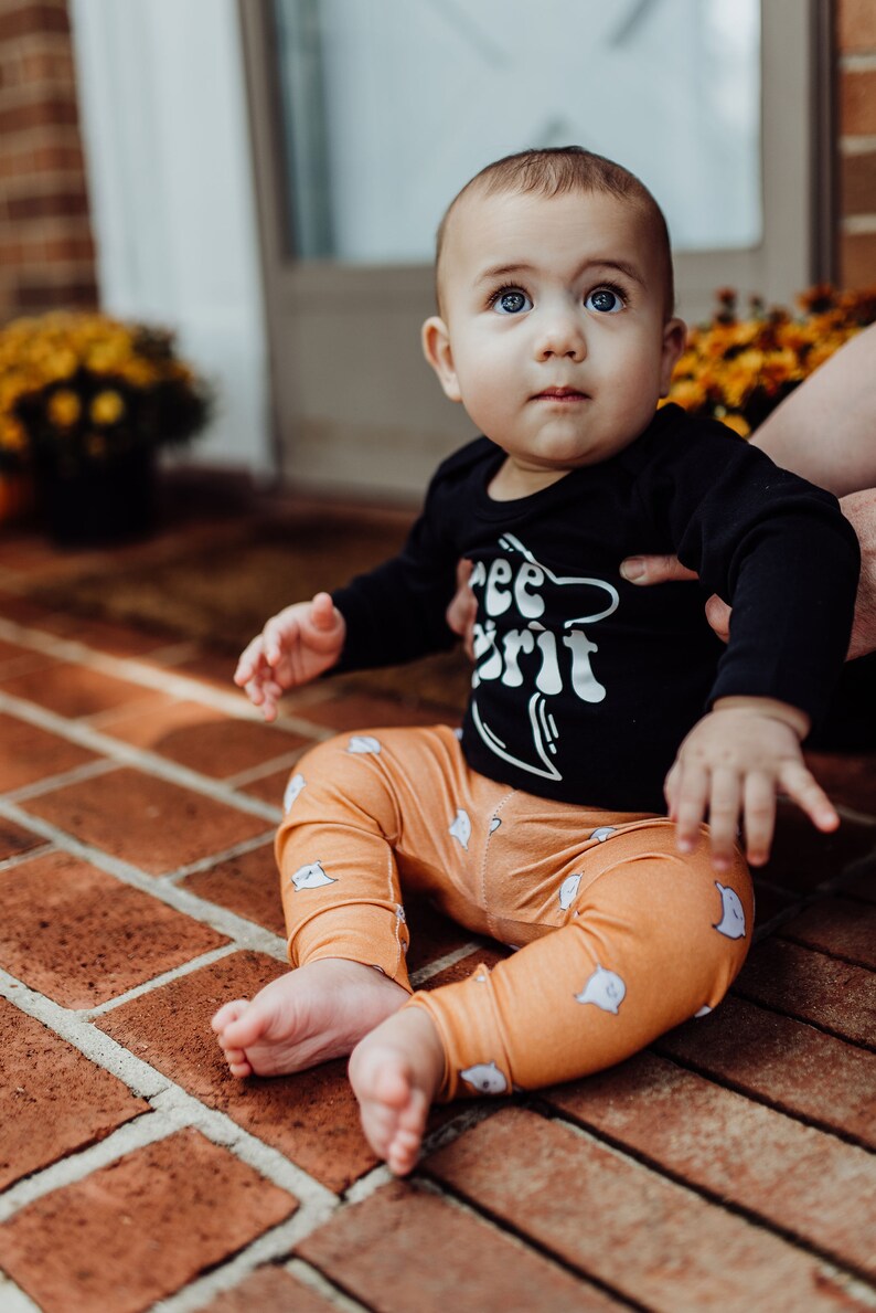 Pu&ograve; includere: Un bambino che indossa una maglietta nera a maniche lunghe con il testo "Free Spirit" e pantaloni arancioni con un motivo a fantasmi. Il bambino &egrave; seduto su un patio in mattoni.