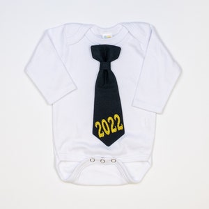 May include: White baby onesie with long sleeves, featuring a black tie with gold glitter text that reads "2022". The onesie has a snap closure at the bottom and is designed for infants.