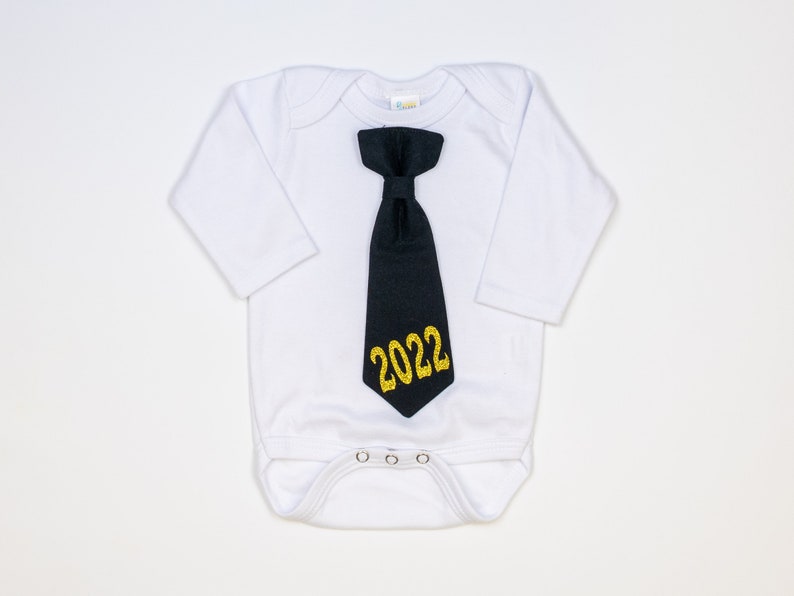 May include: White baby onesie with long sleeves, featuring a black tie with gold glitter text that reads "2022". The onesie has a snap closure at the bottom and is designed for infants.