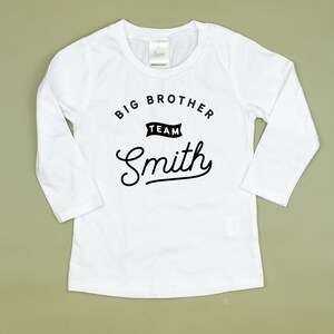 Puede incluir: Camiseta blanca de manga larga con texto negro que dice "Big Brother Team Smith" en una fuente de estilo vintage.