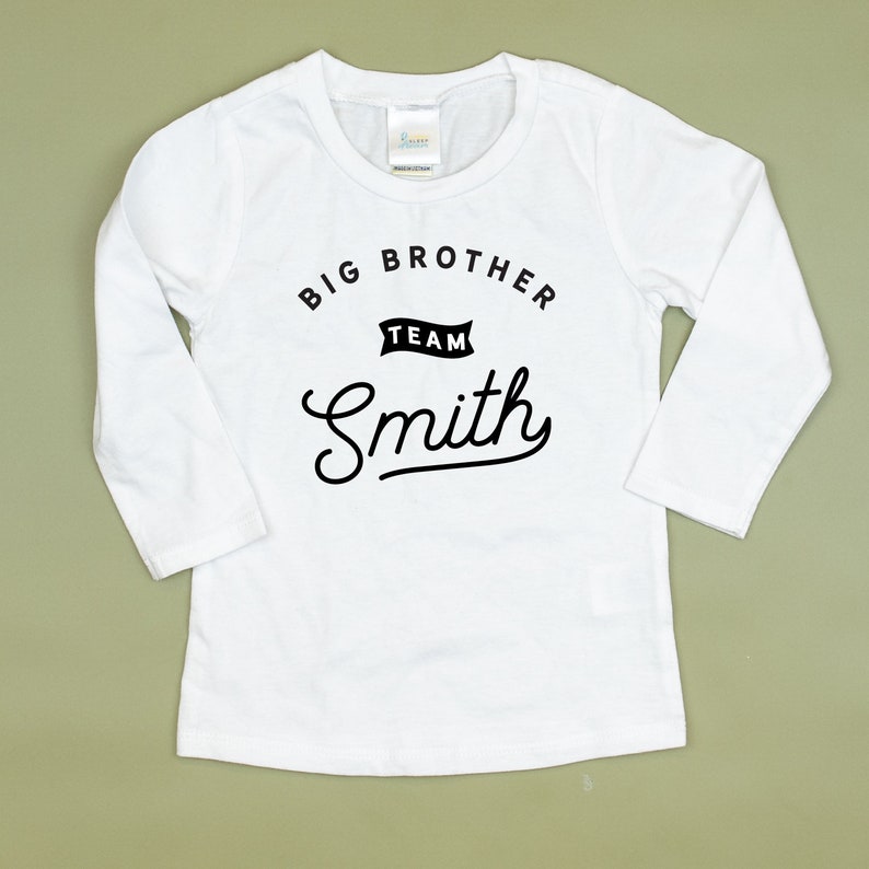 Puede incluir: Camiseta blanca de manga larga con texto negro que dice "Big Brother Team Smith" en una fuente de estilo vintage.