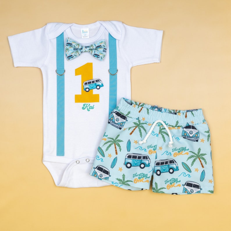 May include: A white baby onesie with blue suspenders and a yellow number one with a blue and white VW bus. The onesie says "Kai" below the number. A pair of blue and white shorts with a VW bus pattern and the words "The Big One" repeated on the shorts.